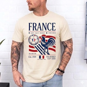 May include: A cream-colored t-shirt featuring a graphic with the text FRANCE, LIBERT&Eacute; &Eacute;GALIT&Eacute; FRATERNIT&Eacute;, Est. 1919, and FIFA WORLD CUP 2026. The design includes a soccer ball, a blue rooster, red and blue stripes, and a small French flag icon.