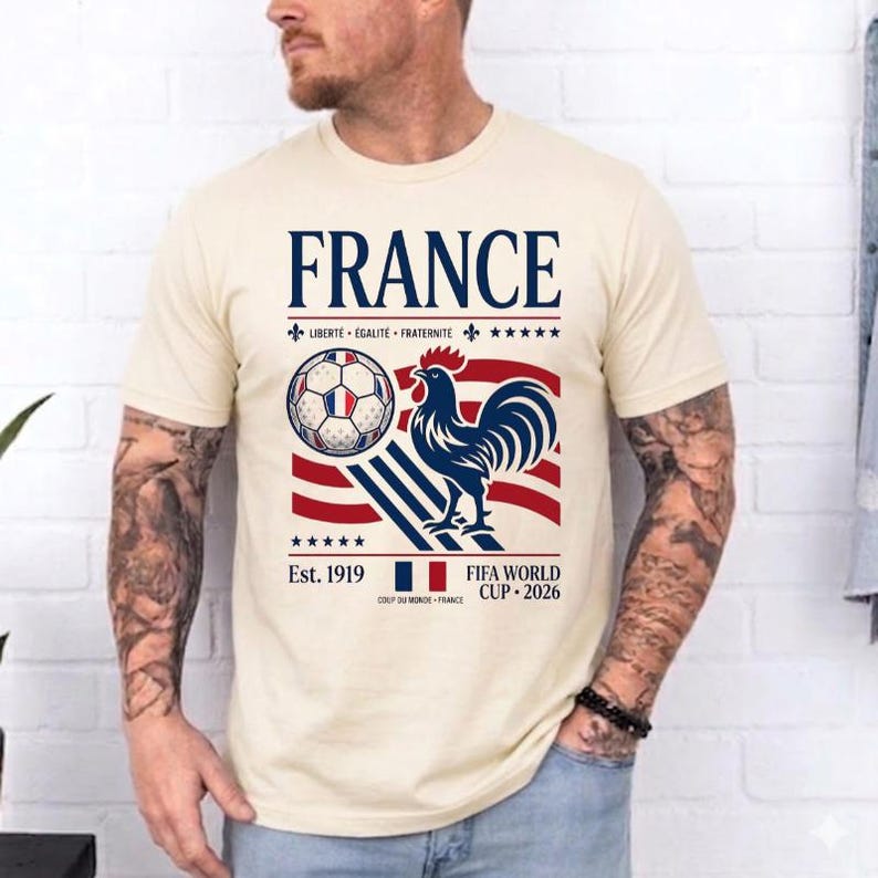May include: A cream-colored t-shirt featuring a graphic with the text FRANCE, LIBERT&Eacute; &Eacute;GALIT&Eacute; FRATERNIT&Eacute;, Est. 1919, and FIFA WORLD CUP 2026. The design includes a soccer ball, a blue rooster, red and blue stripes, and a small French flag icon.