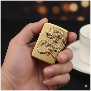 Op de afbeelding: Goudkleurige metalen aansteker met een gedetailleerd drakenontwerp. De draak heeft een rood edelsteen oog en is in reliëf op de voorkant. De aansteker wordt in een hand gehouden, met een vage achtergrond.