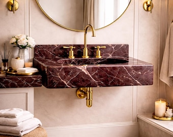 May include: A modern bathroom sink made of burgundy marble with gold fixtures. The sink has a rectangular shape and is mounted on the wall. A gold-framed mirror hangs above the sink, and sconces flank the mirror. A wicker stool holds a stack of white towels.