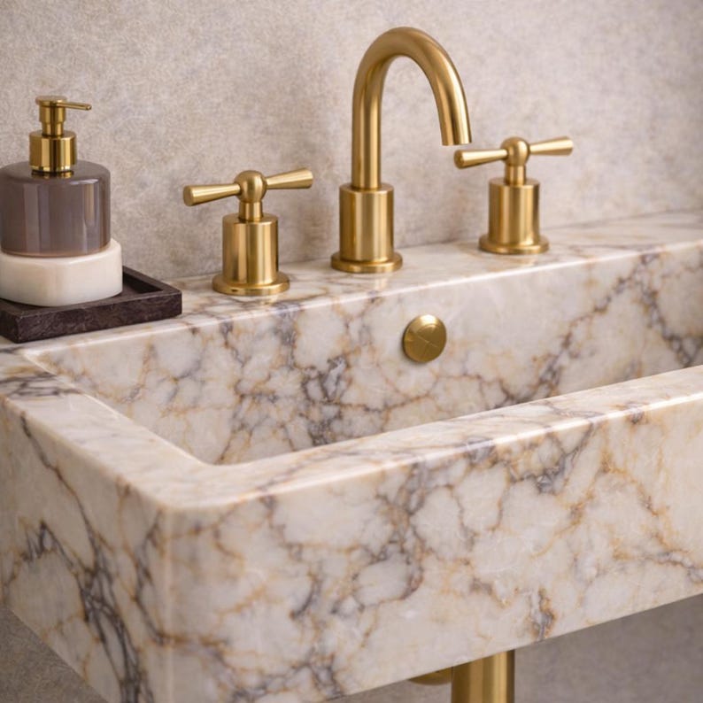 May include: A luxurious bathroom sink with a marble countertop and gold fixtures. The sink has a rectangular basin and a gold faucet with cross-shaped handles. A matching soap dispenser and tray are on the left.
