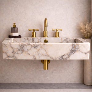 May include: A marble bathroom sink with gold fixtures. The sink has a rectangular basin with a patterned surface. The faucet and handles are gold-colored. A soap dispenser sits on the left side of the sink.