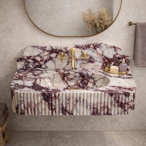 May include: A marble washbasin with a distinctive design. The basin has a white and purple veined pattern, gold taps, and a fluted front. A gold-framed mirror hangs above the basin, with a small tray holding soap and a bottle.