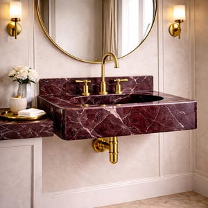 May include: A burgundy marble sink with gold fixtures, including a faucet and two sconces. A round gold mirror hangs above the sink. A small side counter holds a tray with a soap dish and a vase of white flowers.