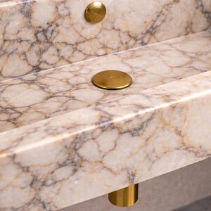 May include: Close-up of a marble sink with a cream and brown veined pattern. The sink has a rectangular basin and is accented with gold-colored drain covers and a gold-colored cylindrical leg.