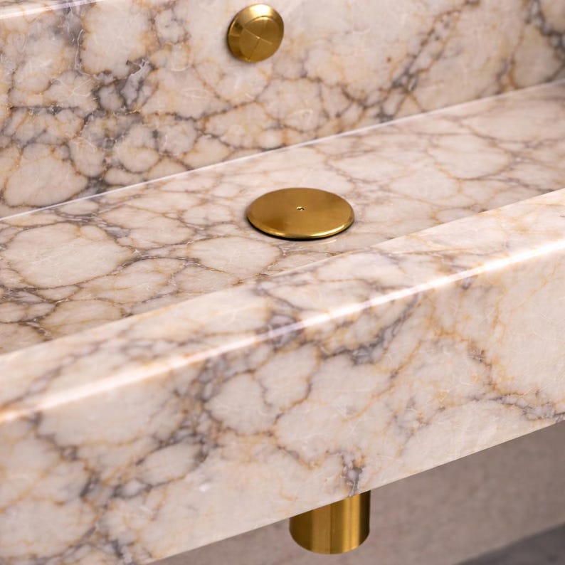 May include: Close-up of a marble sink with a cream and brown veined pattern. The sink has a rectangular basin and is accented with gold-colored drain covers and a gold-colored cylindrical leg.