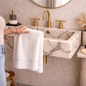 May include: A white, folded towel is held near a marble sink with gold fixtures. The sink has a rectangular basin and is mounted on a wall. A gold faucet and matching handles are visible. A soap dispenser and mirror are also in the background.