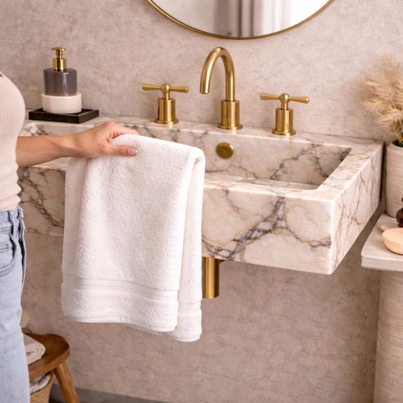 May include: A white, folded towel is held near a marble sink with gold fixtures. The sink has a rectangular basin and is mounted on a wall. A gold faucet and matching handles are visible. A soap dispenser and mirror are also in the background.
