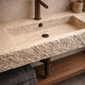 May include: A stone bathroom sink with a bronze faucet and drain. The sink has a rectangular basin and a rough, textured edge. A wooden shelf is below the sink, and a stack of gray towels is on the shelf. The faucet and drain are dark bronze.