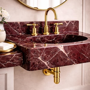 May include: A luxurious bathroom sink with a deep red marble countertop and integrated basin. The sink features gold-colored faucets and drain pipes. A gold-framed mirror is visible in the background, along with a vase of white flowers and a gold tray.
