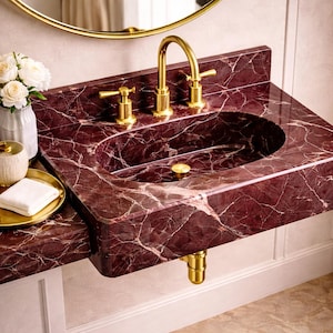 May include: A burgundy marble bathroom sink with gold fixtures. The sink has an oval basin and a matching countertop. A gold faucet and handles are mounted on the countertop. A gold-rimmed mirror is visible in the background.