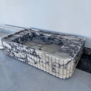 May include: Rectangular marble sink with a white and grey veined pattern. The sink has a fluted exterior design and a central drain hole. The sink is made of stone and is suitable for a bathroom.