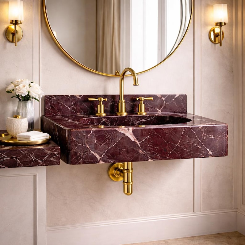 May include: A wall-mounted bathroom sink made of burgundy marble with white veining. The sink has a gold faucet and fixtures. A round gold mirror hangs above the sink, and gold sconces are on either side.