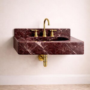 May include: A wall-mounted bathroom sink made of dark red marble with white veining. The sink has a rectangular shape with a built-in basin and is fitted with gold-colored faucets and drain pipes. The sink is mounted on a white wall.