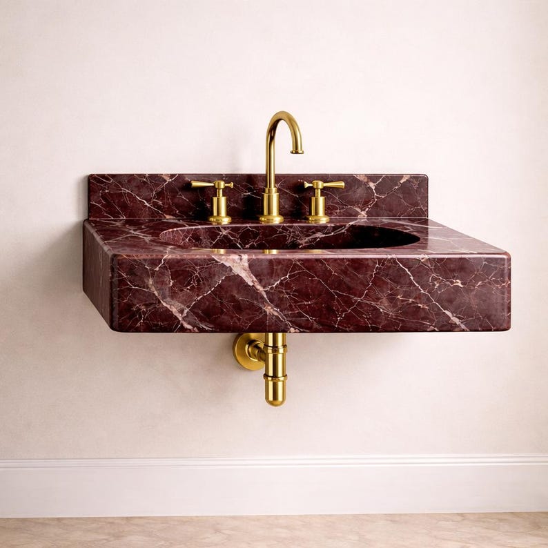 May include: A wall-mounted bathroom sink made of dark red marble with white veining. The sink has a rectangular shape with a built-in basin and is fitted with gold-colored faucets and drain pipes. The sink is mounted on a white wall.