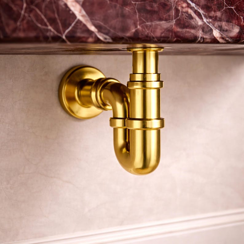 May include: A gold-colored plumbing fixture, likely a P-trap, is mounted on a light-colored wall. The fixture has a curved design with multiple connecting pieces. The top of the fixture is connected to a dark, patterned surface, possibly a countertop.
