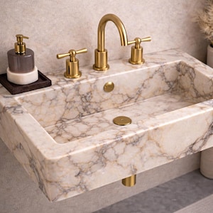 May include: A rectangular marble sink with gold fixtures. The sink has a gold faucet and two handles. A soap dispenser and soap dish sit on the left. The sink is made of beige marble with brown veining.