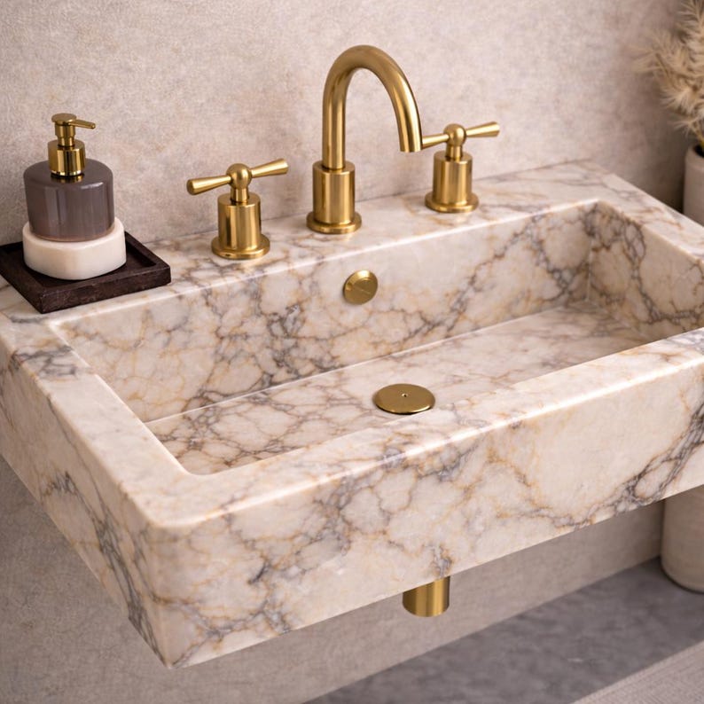 May include: A rectangular marble sink with gold fixtures. The sink has a gold faucet and two handles. A soap dispenser and soap dish sit on the left. The sink is made of beige marble with brown veining.