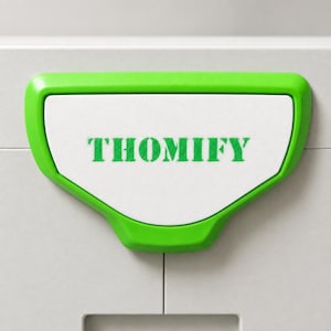 Op de afbeelding: Een felgroene kunststof handgreep met een wit middenpaneel waarop het woord THOMIFY in het groen is gedrukt. De handgreep heeft een trapeziumvorm met afgeronde hoeken en is bevestigd aan een lichtgrijs oppervlak met een zichtbare verticale naad.