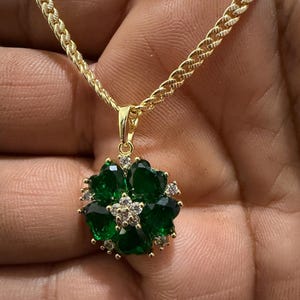 May include: A gold-colored necklace with a flower-shaped pendant. The pendant features dark green gemstones arranged in a floral pattern, with smaller clear stones in the center and around the edges. The chain has a textured, rope-like design.
