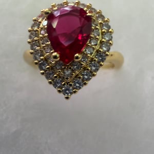 May include: A gold-toned ring featuring a large, pear-shaped, ruby-red gemstone at the center, surrounded by a halo of smaller, clear gemstones. The ring's band is also gold-toned, creating a classic and elegant design.