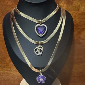 Puede incluir: Tres collares dorados expuestos en un maniquí negro. Cada collar presenta un colgante diferente: un colgante en forma de corazón con una piedra púrpura, un pequeño colgante en forma de corazón con una piedra transparente y un colgante redondo con una piedra púrpura.