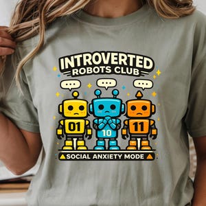 Puede incluir: Camiseta verde salvia con el texto "INTROVERTED ROBOTS CLUB" y tres robots de dibujos animados. Los robots son amarillos, azules y naranjas, cada uno con un número binario. Debajo de los robots está el texto "SOCIAL ANXIETY MODE".