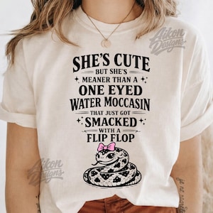 May include: Cream-colored t-shirt with a graphic of a snake wearing a pink bow and the text "She's Cute But She's Meaner Than A One Eyed Water Moccasin That Just Got Smacked With A Flip Flop."