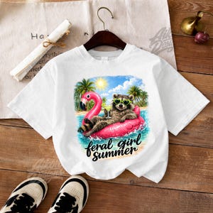 May include: White t-shirt featuring a raccoon wearing heart-shaped sunglasses and relaxing on a pink flamingo float in the water. The graphic includes palm trees, a sun, and the text "feral girl summer".
