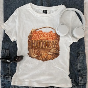 May include: A white t-shirt with a graphic that says "Howdy Honey" in a western-style font. The graphic includes images of cowboy boots, a hat, and roses. The shirt is paired with blue jeans, sunglasses, and white headphones.