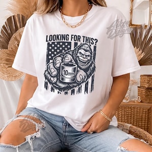 Puede incluir: Camiseta blanca con un gráfico de un Bigfoot sonriente sosteniendo una llave de vaso de 10 mm frente a una bandera estadounidense. El texto "LOOKING FOR THIS?" está encima del gráfico. La camiseta es de estilo casual.
