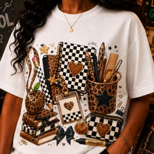 May include: A white t-shirt featuring a graphic print of school supplies including a leopard print pencil cup, a checkered notebook with a heart, a leopard print apple, a stack of books, a checkered pencil case, and a highlighter labeled HIGHLIGHTER.