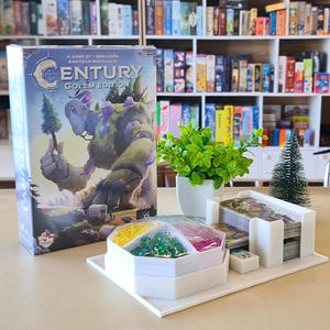 Puede incluir: Caja del juego de mesa "Century: Golem Edition" con una ilustración de un golem. Un organizador blanco contiene piezas y cartas del juego. Una pequeña planta verde y un árbol de Navidad en miniatura también están sobre la mesa.