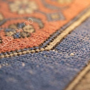 Op de afbeelding: Close-up van een deel van een tapijt met een gedetailleerd patroon. Het tapijt heeft een combinatie van kleuren, waaronder tinten blauw, oranje en beige. Het ontwerp bevat bloem- en geometrische elementen, met zichtbare textuur.