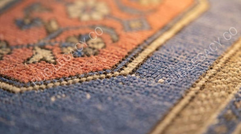 Op de afbeelding: Close-up van een deel van een tapijt met een gedetailleerd patroon. Het tapijt heeft een combinatie van kleuren, waaronder tinten blauw, oranje en beige. Het ontwerp bevat bloem- en geometrische elementen, met zichtbare textuur.