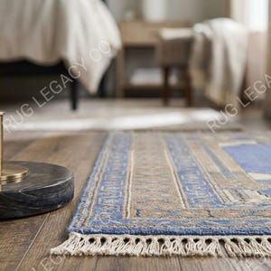 Op de afbeelding: Een rechthoekig blauw en beige tapijt met een gedetailleerde rand en franjes. Het tapijt heeft een centraal ontwerp met geometrische patronen en ligt op een houten vloer. Het tapijt maakt deel uit van een woondecoratiecollectie.