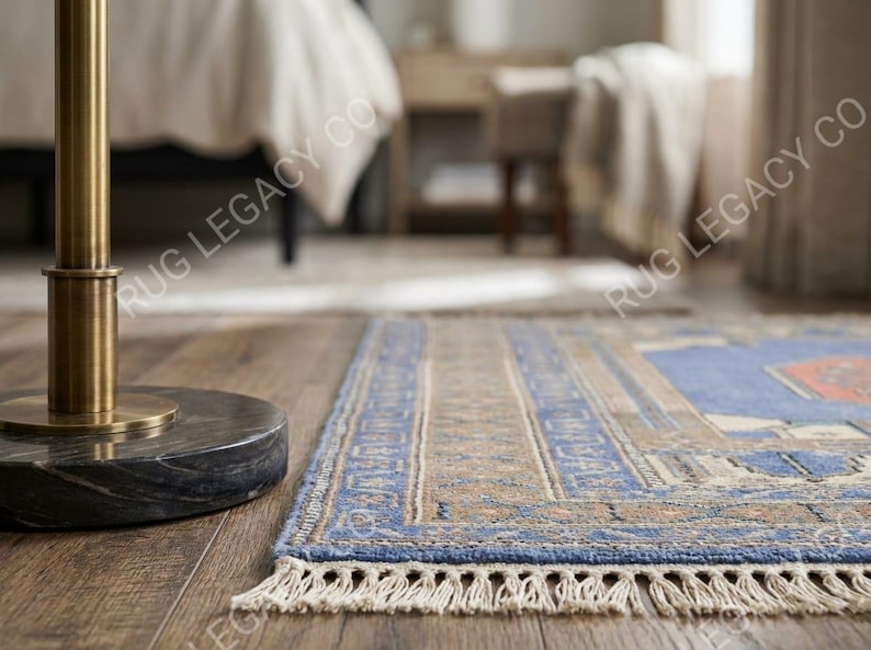 Op de afbeelding: Een rechthoekig blauw en beige tapijt met een gedetailleerde rand en franjes. Het tapijt heeft een centraal ontwerp met geometrische patronen en ligt op een houten vloer. Het tapijt maakt deel uit van een woondecoratiecollectie.