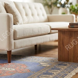 Op de afbeelding: Een cr&egrave;mekleurige bank met knopen en houten poten in een lichte kamer. Een houten salontafel met boeken en een kaars. Een patroonkleed in blauwe en rode tinten ligt op de vloer.