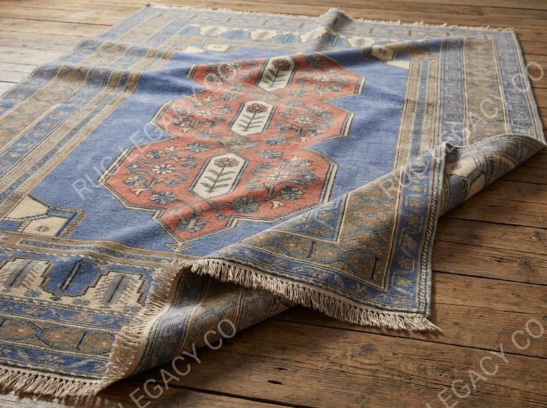 Op de afbeelding: Een rechthoekig tapijt met een blauw veld en een centraal koraalkleurig medaillon met bloemmotieven. Het tapijt heeft een rand met geometrische patronen en franjes langs de randen. Het tapijt ligt op een houten oppervlak.