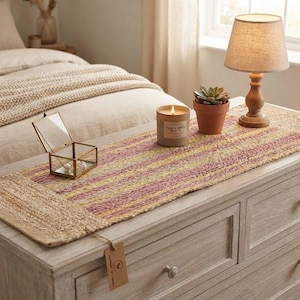 May include: A rectangular woven table runner featuring alternating horizontal stripes of natural tan, muted pink, and soft yellow. The runner is placed on a wooden dresser and includes a small brown paper tag attached to one corner with a thin string.