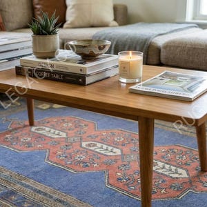 Op de afbeelding: Een houten salontafel in mid-century modern stijl met een rechthoekig blad en schuine poten. De tafel bevat boeken, een kaars, een kom en een kleine plant in een pot. Onder de tafel ligt een patroonkleed in blauw en rood.