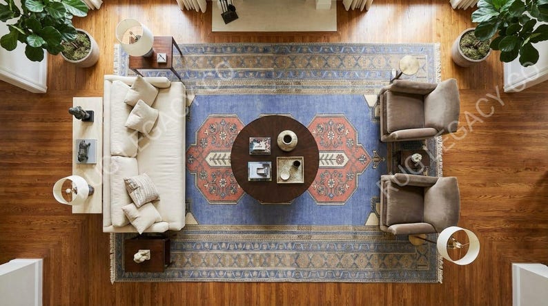 Op de afbeelding: Luchtfoto van een woonkamer met parketvloer, een blauw patroonkleed en een ronde salontafel. Een cr&egrave;mekleurige bank met kussens, twee fauteuils en lampen zijn rond het kleed geplaatst. Potplanten voegen groen toe.