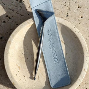 May include: A silver-colored facial tool with a curved design, resting in a light blue case. The case is embossed with "BREAKING WAVE". The tool and case are displayed on a beige, textured surface, suggesting a skincare or beauty product.