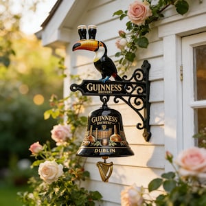 May include: A black Guinness Brewery bell and sign with a toucan figure. The bell has a gold harp and the sign reads "Guinness Brewery Dublin". The toucan has two pints of Guinness on its head. The sign is mounted on a white wall with pink roses.