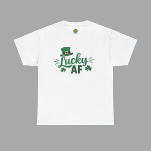 Könnte beinhalten: Weißes T-Shirt mit einem grünen Koboldhut und den Worten "Lucky AF" in Grün. Das Design beinhaltet Kleeblätter und dekorative Linien. Ein festliches T-Shirt für St. Patrick's Day.