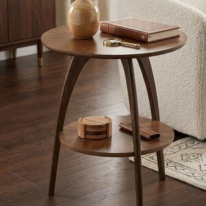 May include: A dark brown, round side table with a lower shelf. A brown book, a magnifying glass, and a decorative vase sit on the top. A set of coasters and a remote are on the lower shelf. The table has three curved legs.