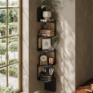 May include: A black corner shelf unit with a woven cane backing. The shelves display a potted plant, books, a framed picture, a clock, and decorative items. The unit is against a neutral wall near a window.