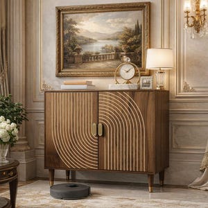 May include: A wooden cabinet with a decorative gold pattern on the doors. The cabinet has gold handles and legs. A painting hangs above the cabinet, and a lamp and clock sit on top. A robotic vacuum cleaner is on the floor.