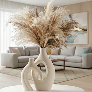 May include: Two beige ceramic vases with unique, curved designs sit on a white table. The larger vase has a circular opening. Both vases hold a large arrangement of dried pampas grass and other dried flowers, creating a neutral, natural aesthetic.