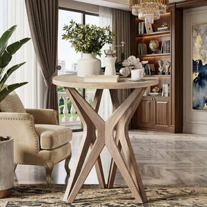 May include: A light-colored wooden side table with a round top and a unique, geometric base. The table is in a well-lit room with a neutral color palette. A white vase with greenery, a coffee grinder, and a cup and saucer are on the table.